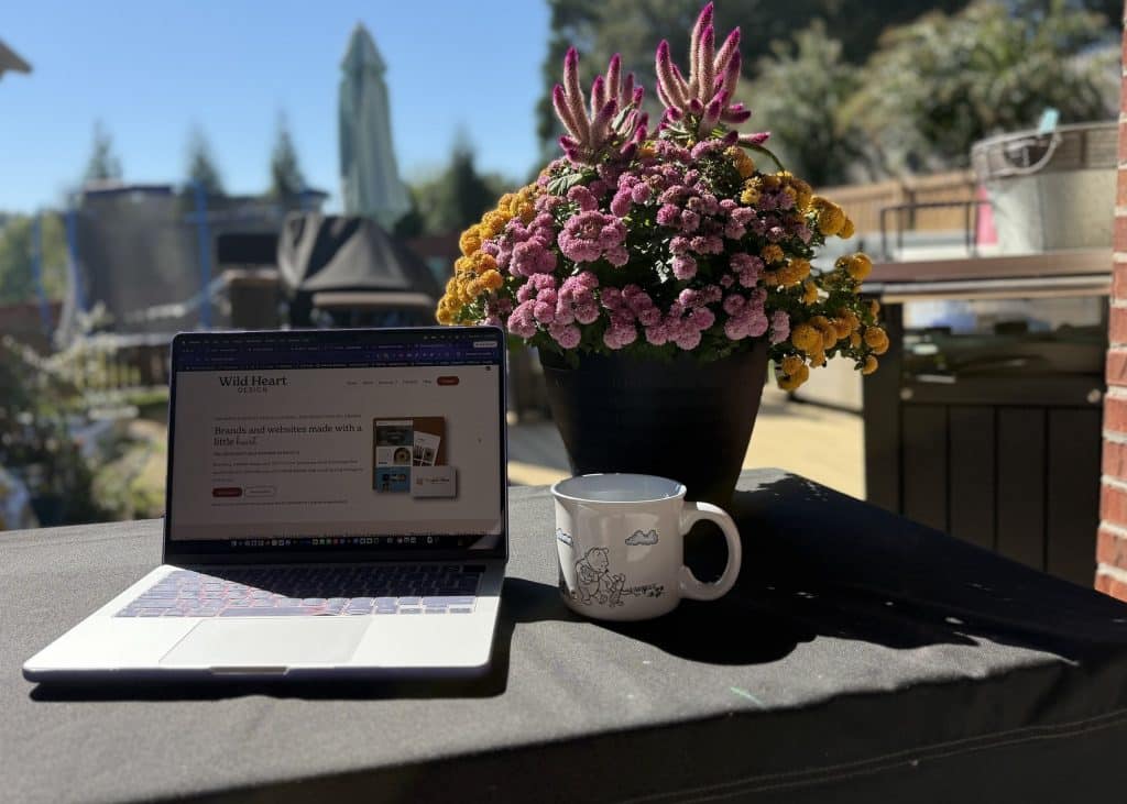 Laptop showing the Wild Heart Design website on an outdoor table beside a pot of fall mums and a coffee mug in afternoon sun
